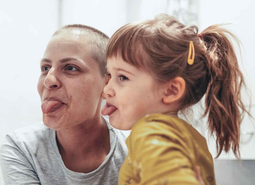 Frau mit Krebserkrankung und ihre Tochter strecken die Zunge raus und habe Spaß.