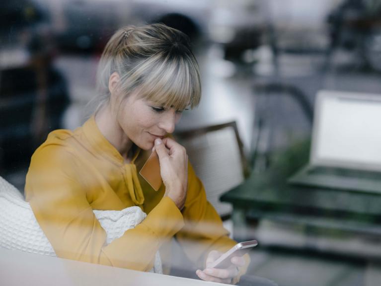 Blonde Frau mit gelber Bluse sitzt an einem Schreibtisch und schaut in ihr Smartphone.