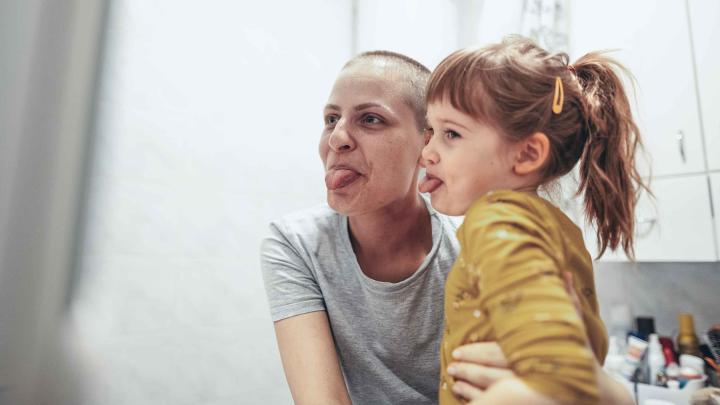 Krebserkrankte Mutter sitzt mit ihrer Tochter vor dem Spiegel und streckt die Zunge raus