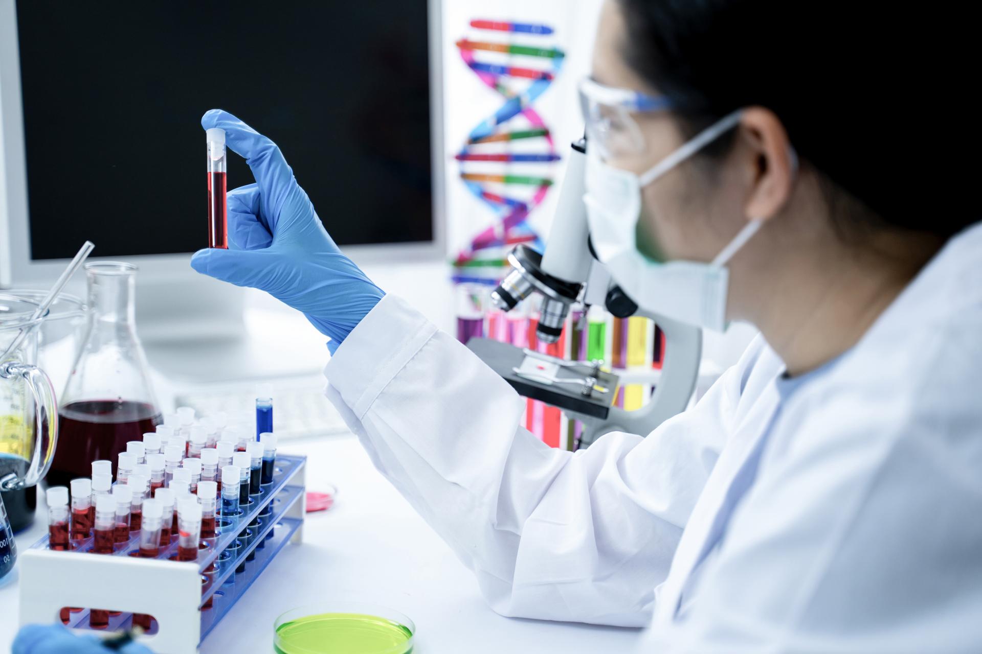 Eine Frau sitzt im Labor, trägt einen weißen Kittel, Handschuhe und einen Mundschutz. Sie untersucht eine Blutprobe auf DNA Auffälligkeiten.