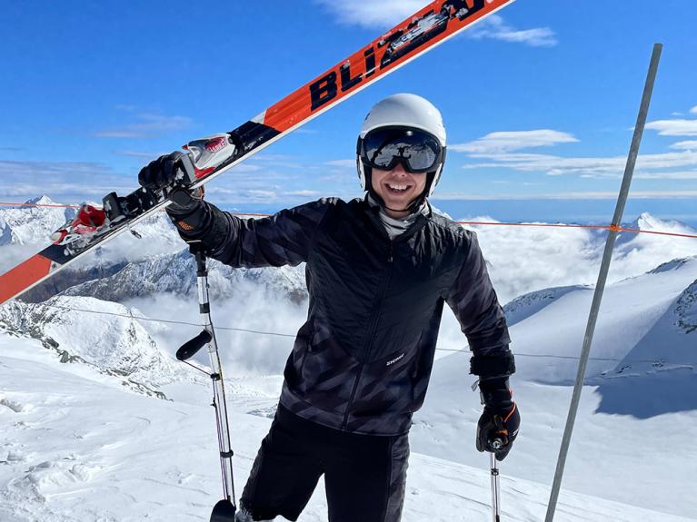 Amputierter Skifahrer mit schwarzer Jacke hebt glücklich seine Ski in die Luft und lacht.