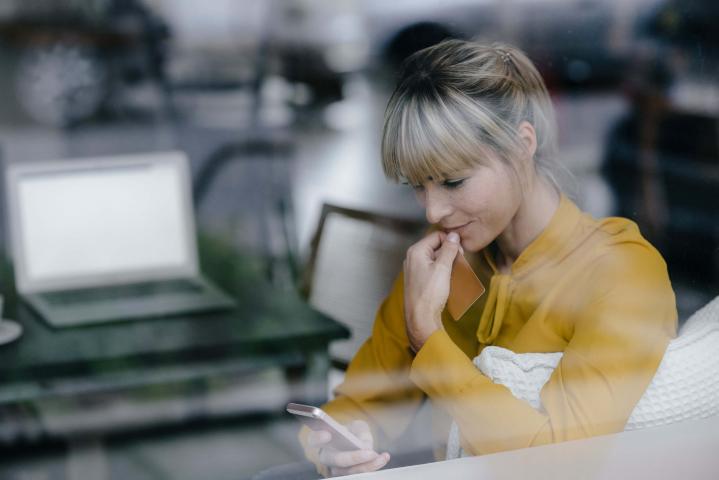 Blonde Frau mit gelber Bluse sitzt hinter einer Glasscheibe und betrachtet Informationen auf ihrem Smartphone.