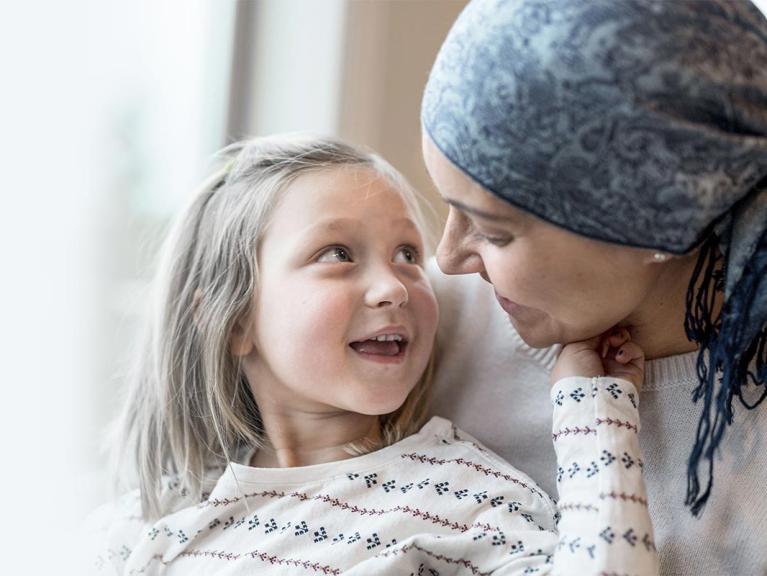 Krebskranke Mutter sitzt mit ihrer Tochter am Fenster und lächelt