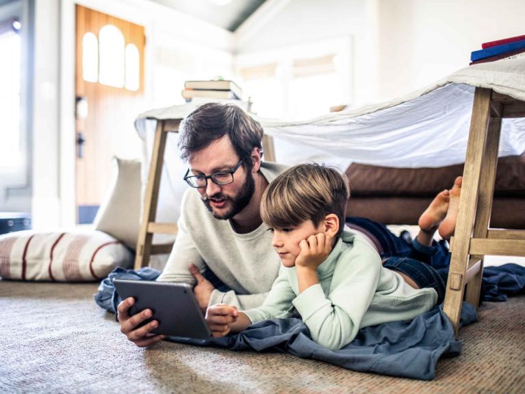Ein junger Vater schaut sich gemeinsam mit seinem Sohn ein Video auf dem Tablet an und liegt im Kinderzimmer auf dem Boden