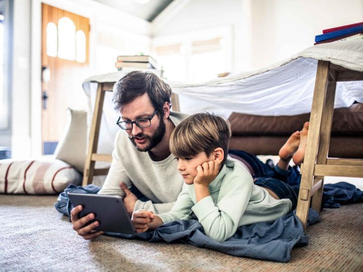 Ein junger Vater schaut sich gemeinsam mit seinem Sohn ein Video auf dem Tablet an und liegt im Kinderzimmer auf dem Boden