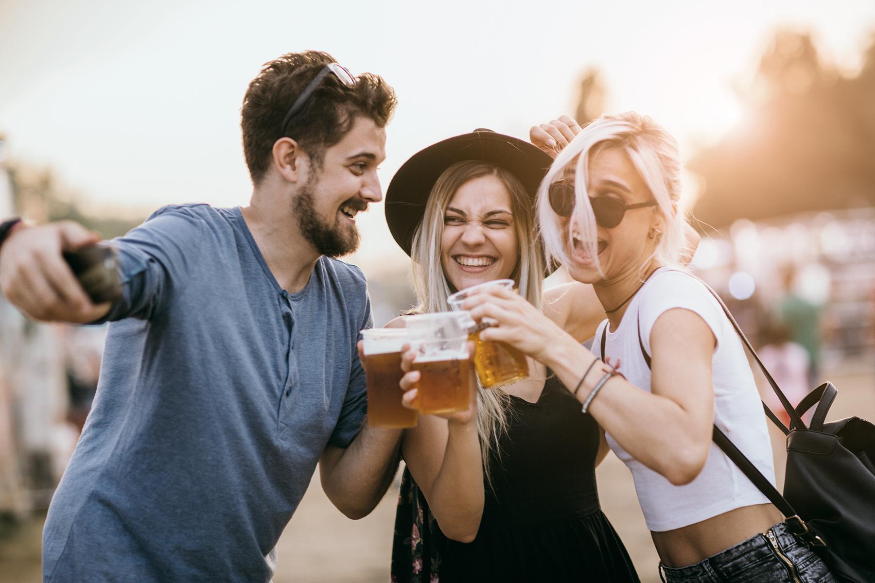 2 junge Frauen und ein junger Mann stoßen lächelnd mit Alkohol an