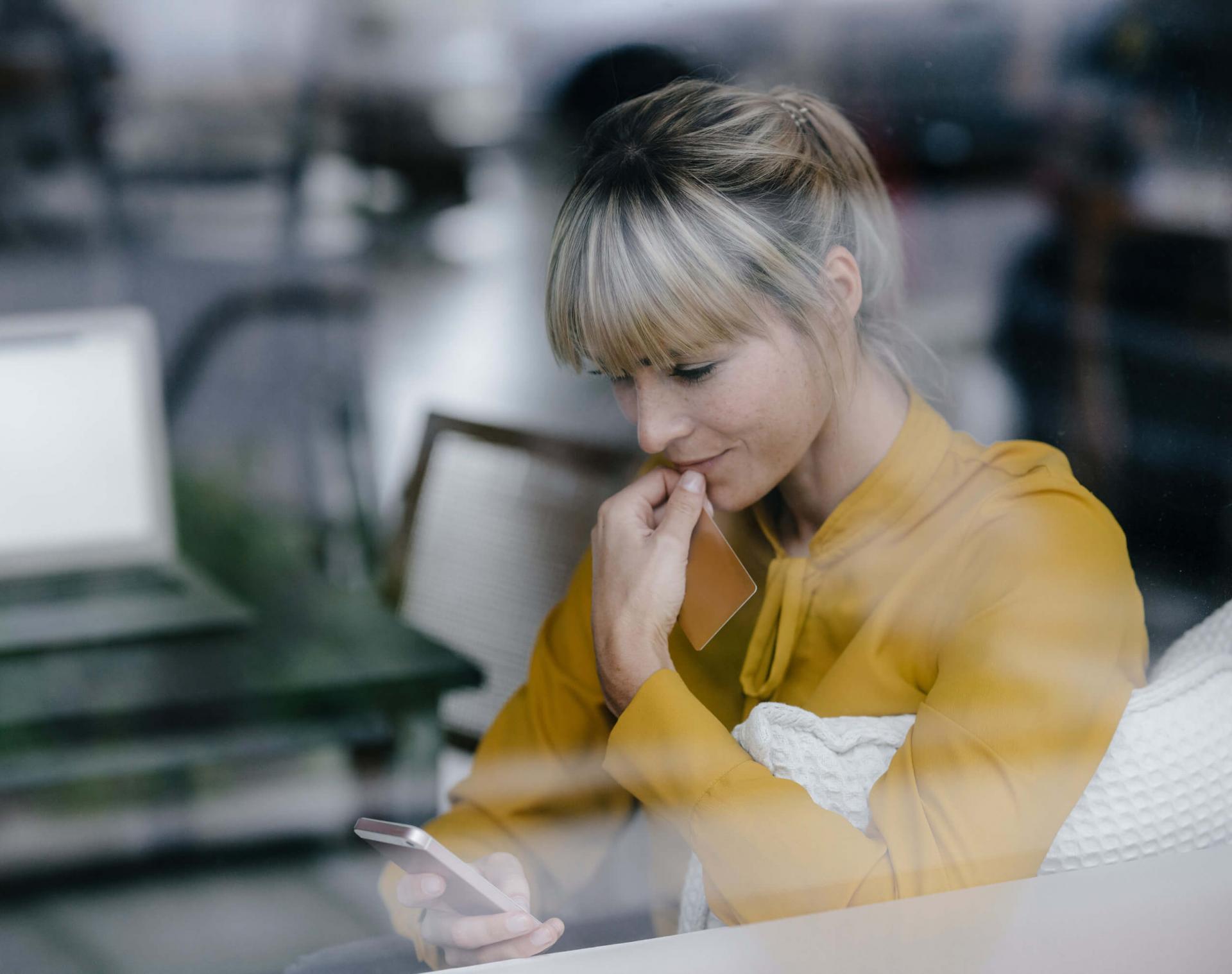 Blonde Frau mit gelber Bluse sitzt hinter einer Glasscheibe und betrachtet Informationen auf ihrem Smartphone.