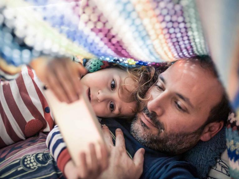 Vater liegt mit seiner Tochter unter einer Decke und liest etwas vor.