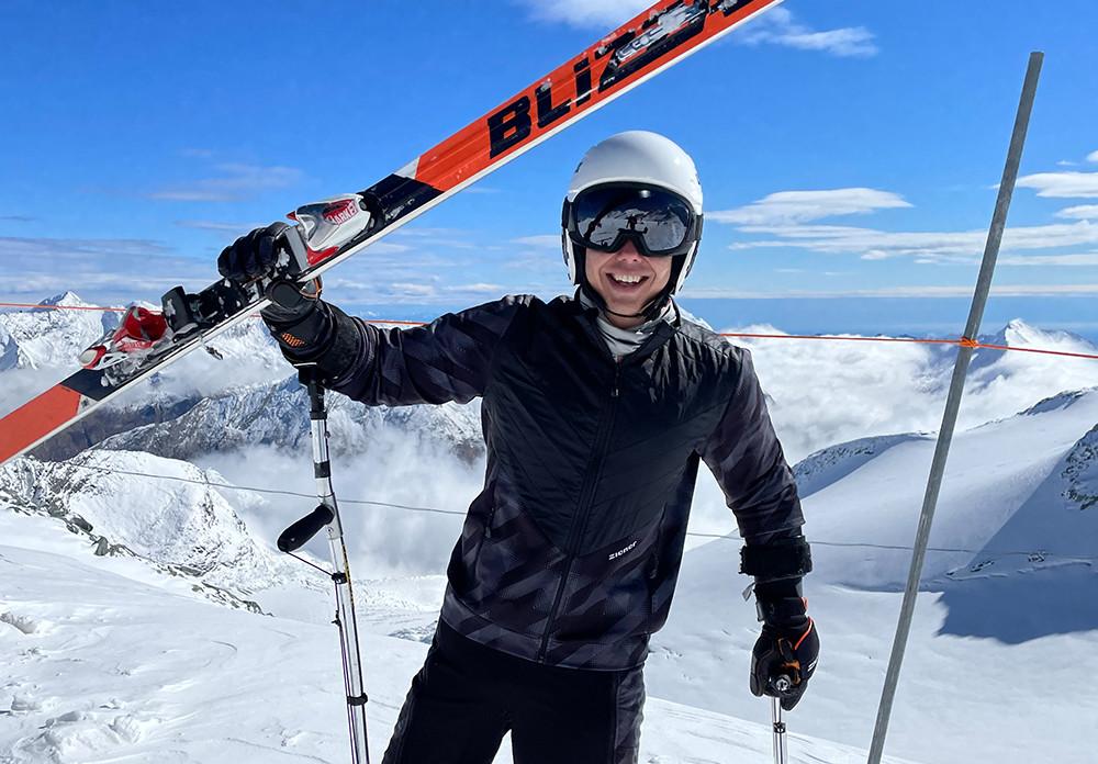 Amputierter Skifahrer mit schwarzer Jacke hebt glücklich seine Ski in die Luft und lacht.