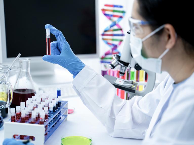 Eine Frau sitzt im Labor, trägt einen weißen Kittel, Handschuhe und einen Mundschutz. Sie untersucht eine Blutprobe auf DNA Auffälligkeiten.
