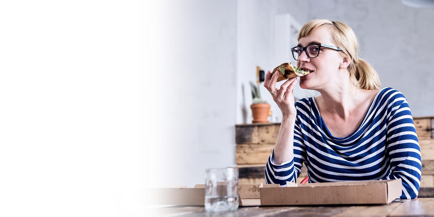 Eine blonde Frau mit schwarzer Brille sitzt am Esstisch und isst ein Stück Pizza.