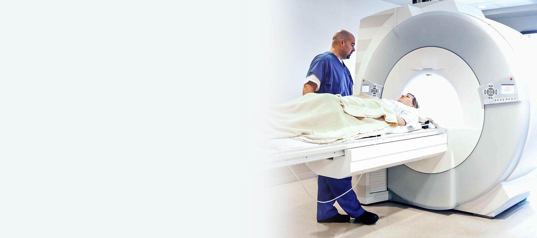Der Pfleger trägt einen blauen Arbeitskittel und schiebt den Patienten liegend in die MRT-Röhre