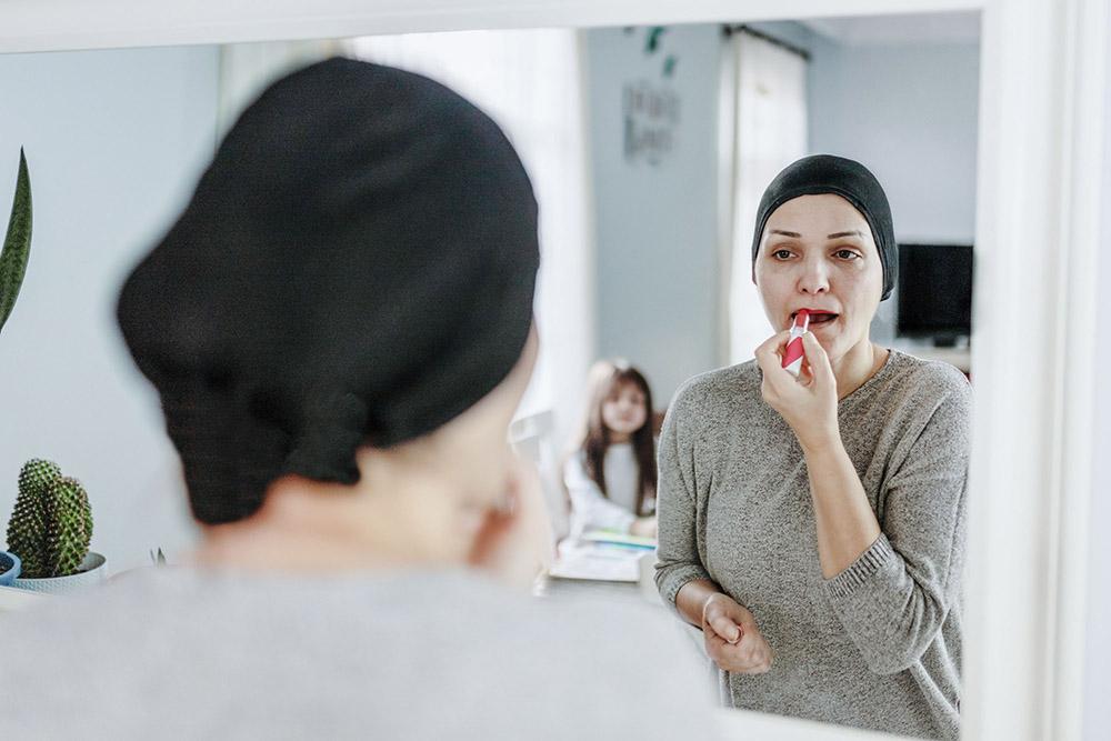 Krebserkrankte Frau mit Kopftuch steht vor dem Spiegel und schminkt sich