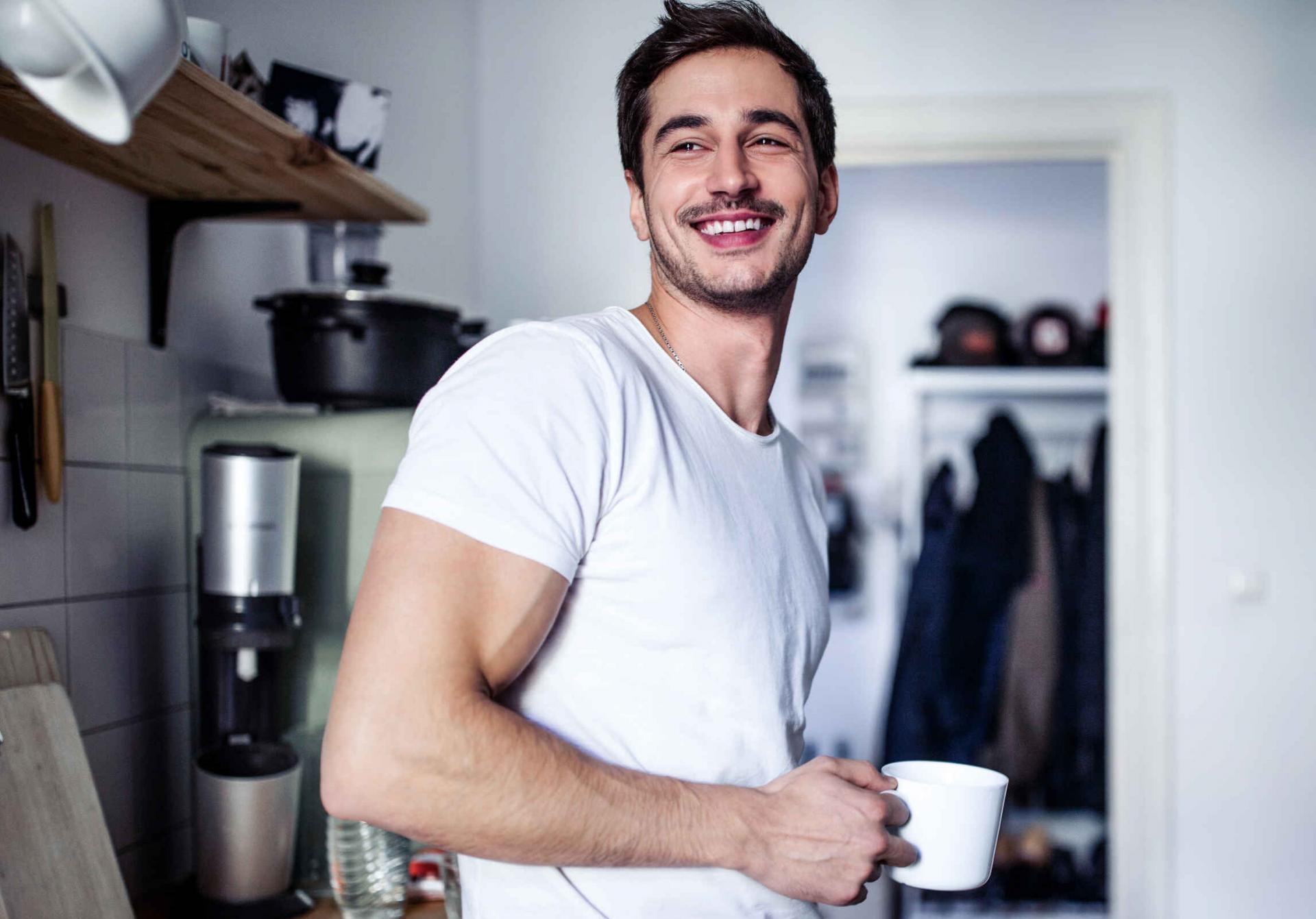 Junger Mann im weißen T-Shirt lehnt lachend in der Küche und hält einen weißen Becher in der Hand