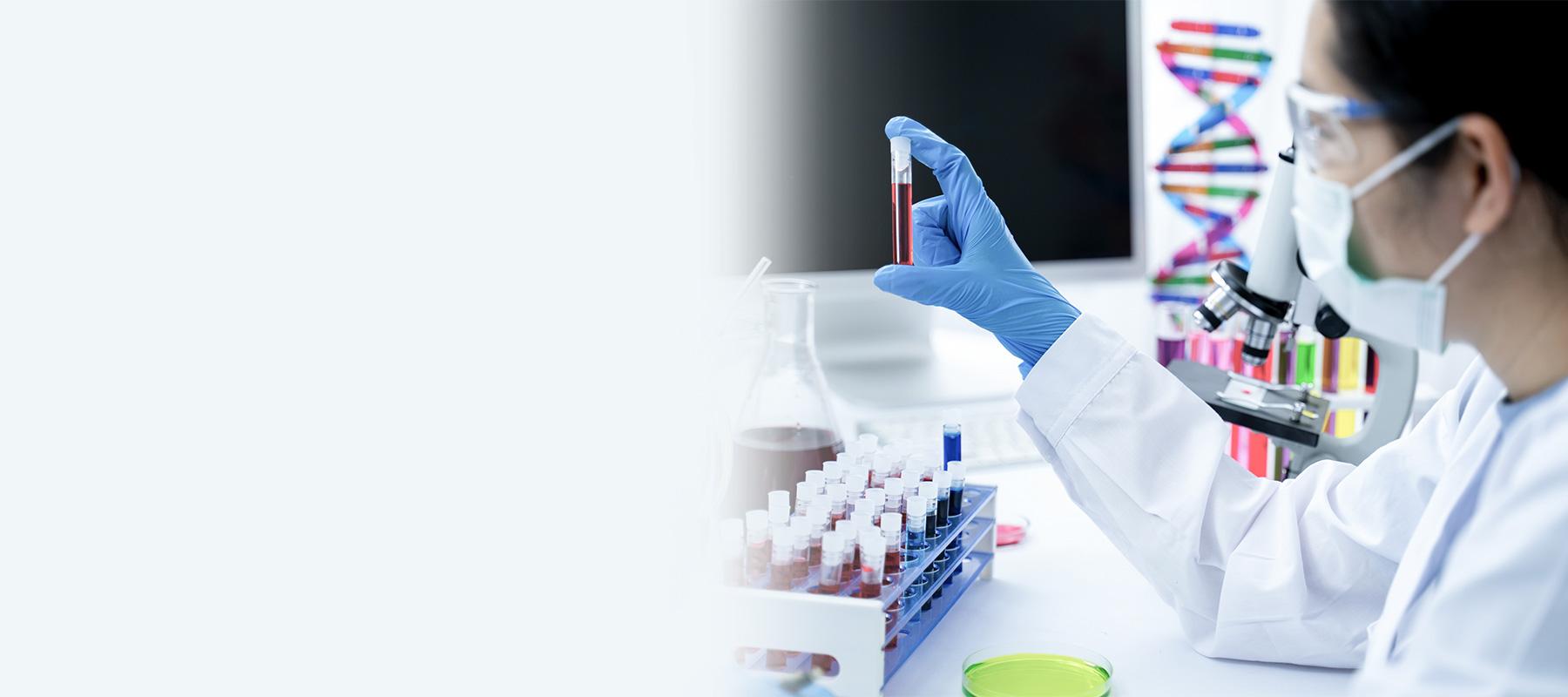 Eine Frau sitzt im Labor, trägt einen weißen Kittel, Handschuhe und einen Mundschutz. Sie untersucht eine Blutprobe auf DNA Auffälligkeiten..