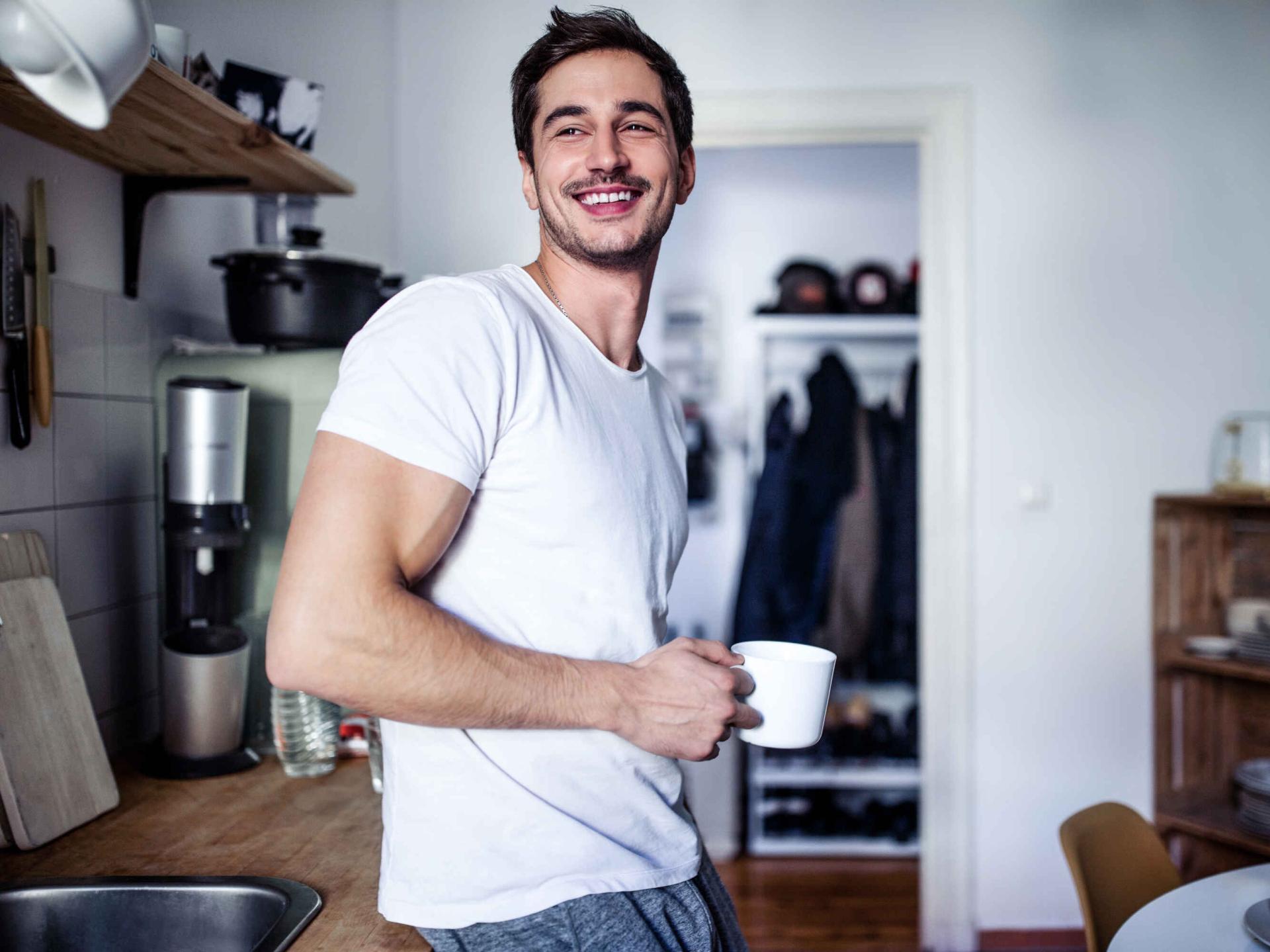 Mann steht glücklich mit einer Kaffeetasse in der Küche.