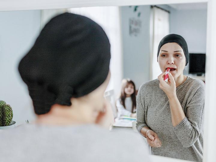 Krebserkrankte Frau mit Kopftuch steht vor dem Spiegel und schminkt sich
