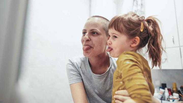 Eine krebskranke Mutter sitzt mit ihrem Tochter im Bad vor dem Spiegel und beide strecken ihre Zunge raus.