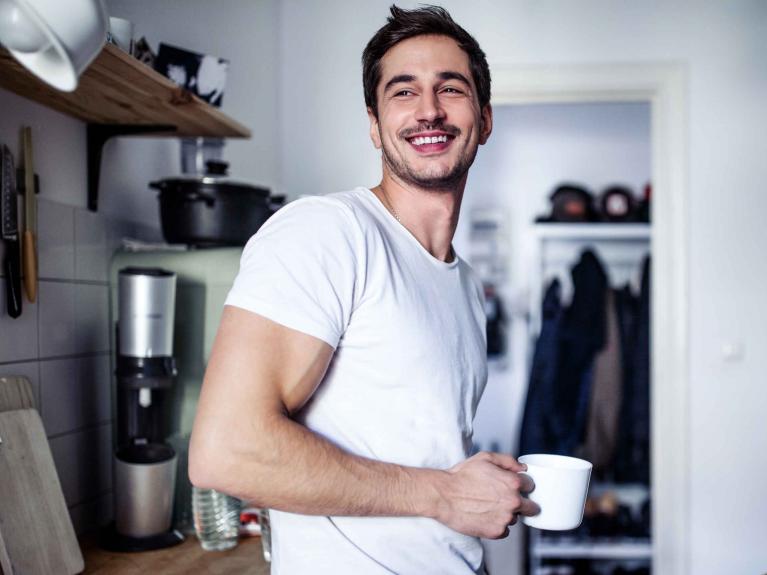 Junger Mann im weißen T-Shirt lehnt lachend in der Küche und hält einen weißen Becher in der Hand